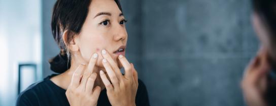 Woman examining acne in mirror and squeezing blemish, illustrating difference between acne and pimples for skincare and dermatology blog.
