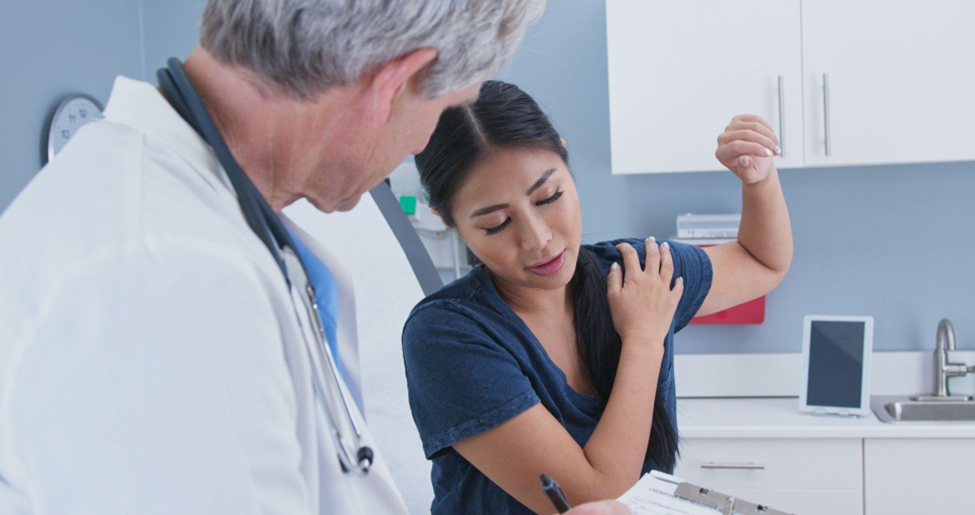 Japanese woman explaining shoulder pain to doctor in exam room.